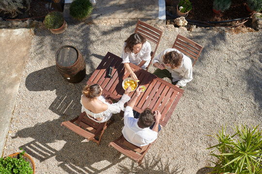 A top-down view of friends enjoying a sunny outdoor meal at a rustic wooden table, surrounded by a quaint garden setting, epitomizing relaxed summer dining.