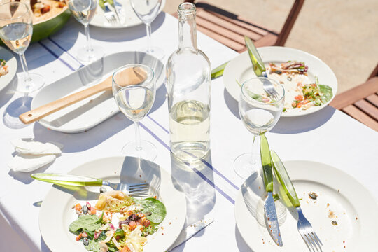 The remains of a summer feast lay on a sunlit table, with empty dishes, scattered crumbs, and half-empty glasses of white wine, capturing the relaxed end of a joyful meal.