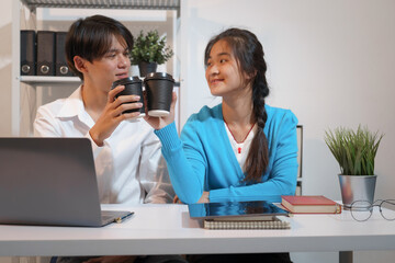 An Asian male consultant and an African American female intern sit at a table with laptops doing...