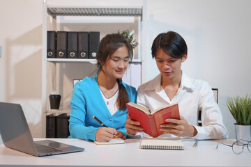 An Asian male consultant and an African American female intern sit at a table with laptops doing...