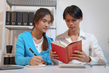 An Asian male consultant and an African American female intern sit at a table with laptops doing...