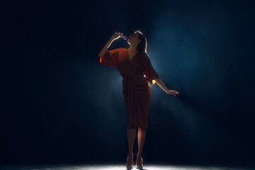 Female silhouette, singer under glowing bright spotlights at stage in smoke against black...