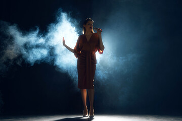 From shadows, voice takes flight. Woman singing into microphone surrounded dramatic smoke and backlighting on stage against black background. Performance. Concept of hobby, festival, concert, disco. - obrazy, fototapety, plakaty
