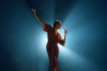 Graceful singer with uplifted arm, bathed in serene blue spotlight on stage against dark background...
