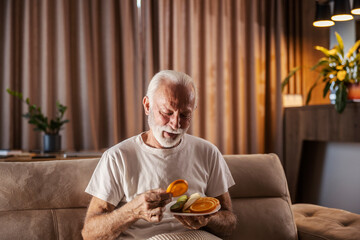 An ill senior man is eating fruits full of vitamins while sitting at home.