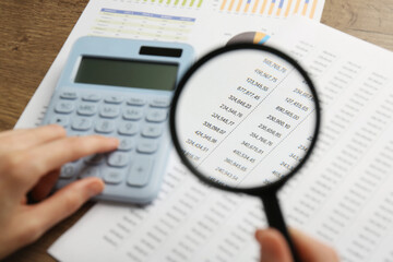 Woman looking at accounting document through magnifying glass while using calculator at wooden...