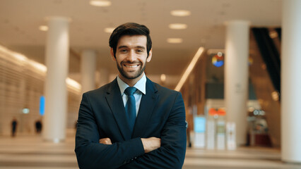 Professional business man standing with arm folded at modern mall. Smiling caucasian investor looking at camera with confident while crossing arms at hall with blurring background. Portrait. Exultant.