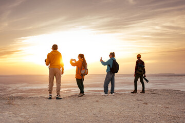 Four young hikers and photographers with backpacks are standing at sunset view point and taking...