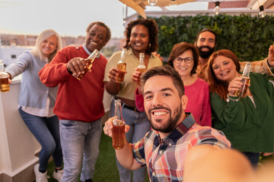 Group selfie of multiethnic friends on rooftop terrace toasting with beer at sunset