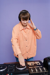 Cool young boy DJ mixes music on a mixing console  in stylish clothes on a color background.