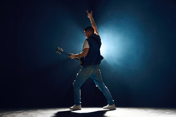 Rear view. Guitar player engaging with music and show Rock-n-roll gesture. Vocalist highlighted by...