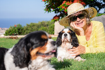 Happy senior woman with hat and sunglasses lying outdoors in the park with two cavalier king charles spaniel dogs. Retired elderly lady enjoying her best friends