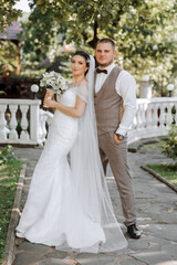 Naklejka premium A handsome groom embraces his bride in a lush white dress and smiles in a beautiful outdoor setting. Under the open sky. High quality photo. A newlywed couple poses together on a sunny summer day.