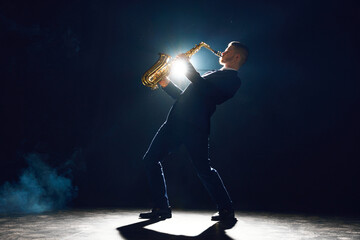 Elegant man with sax tilted upwards performing new melody on stage against dark background with...