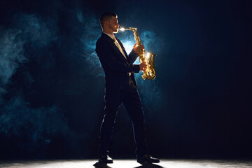 Solo saxophonist in concert outfit performing with intense expression on stage with backlight against dark background with dramatic smoke. Concept of art, instrumental music, dance, culture. Ad - obrazy, fototapety, plakaty