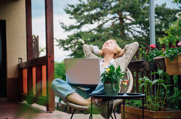 Fototapeta na wymiar Young blonde woman in her country house is sitting on the terrace surrounded by flowers and using a laptop. Relaxed female freelancer enjoying her life.