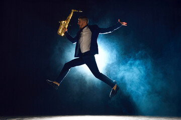 Dynamic shot of artistic man, solo performer jumping while playing saxophone on stage with...