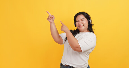 A chubby Asian woman wearing a headphones is pointing at something on a yellow background.