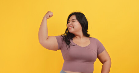 A chubby Asian woman wearing exercise clothes is showing off her physical strength on a yellow background.