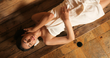 Young woman in towel relaxing in wooden sauna at spa. Asian woman in bathrobe doing body treatment...