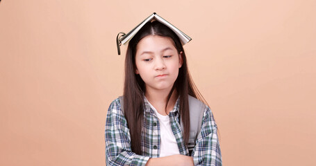 Cute school girl is carrying a school bag and holding a notebook on her head. Isolated on brown background in studio.