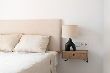 Part of a cozy large bed in a bedroom with beige bed linen and a lamp on bedside table next to it. Scandinavian interior concept in a new building for young people