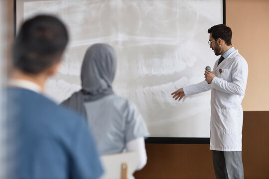 Medium full shot of Muslim man medic holding microphone showing dental X-Ray on projection screen to audience during medical conference
