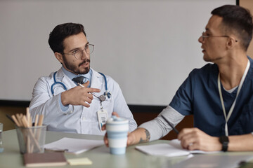 Medium shot with focus on young Muslim man doctor in glasses talking with defocused male colleague sitting at desk in meeting room