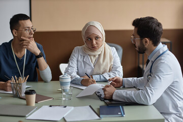 Muslim woman doctor in beige headscarf carefully listening to male colleague and making notes while...