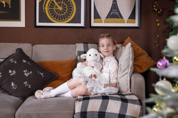 A little blonde girl sits on a sofa in a cozy living room at home and hugs a soft toy. Photos in the interior with a New Year tree. The concept of joy and happy childhood, holiday