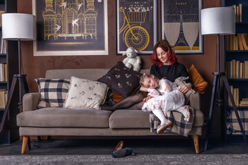 A little girl and her mother are sitting hugging each other on the sofa in a cozy living room at home. Photos in the interior. Concept of joy and happy childhood, holiday