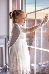 A little angel stands on the windowsill and looks out the window. Cute little angel girl looks and...