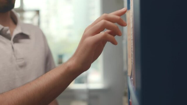 Close-up shot of hand and face of young Middle Eastern man standing in front of bookcase in public library, touching spines of books while reading names to choose material for thesis