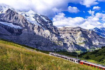 Idyllic swiss landscape scenery. Green pastures, snowy peaks of Alps mountains and railway road with passing train. Kleine Scheidegg station, Switzerland. - 723812651
