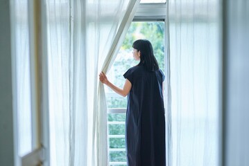 レースカーテンを開けて窓から外を見る若い女性