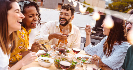 Multiethnic friends having fun at rooftop bbq dinner party - Group of young people diner together sitting at restaurant dining table - Cheerful multiracial teens eating food and drinking wine outside - obrazy, fototapety, plakaty