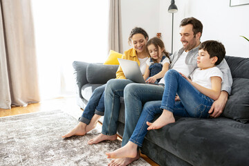 Happy family of four using laptop sitting on the sofa in cozy apartment, parents and two kids...