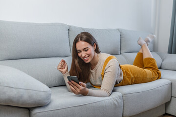 Smiling woman lying on sofa celebrating with clenched fist using cell phone
