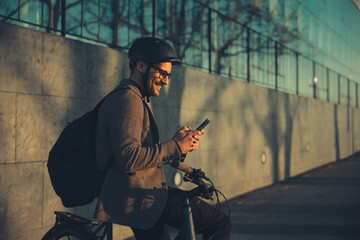 A profile of a smiling young adult businessman with a backpack typing a text message and leaning on...