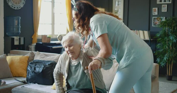Nurse assisting senior grandmother in walking at home. Old woman has a problem with her legs and it is difficult to walk, she feels faint while standing up with the help of a caregiver