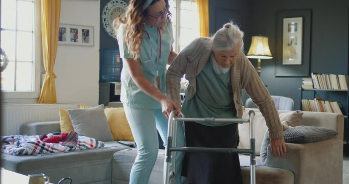 Nurse caregiver helping senior woman getting up from sofa and walk with a walker.