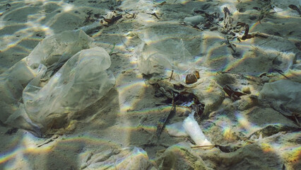 Plastic garbage lies on sandy-silty bottom of sea in shallow water on bright sunny day in sun glare. Seabed polluted with plastic and other debris, Red sea, Safaga, Egypt