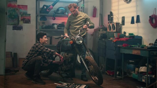 Background of home garage. Boy in casual clothes repairing old retro motorcycle. Father and son spending time together inspecting motorbike. Maintenance service and small family business concept.