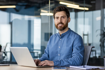 Portrait of a successful and confident young man in a blue shirt sitting in the office at a desk, working on a laptop and smiling at the camera - obrazy, fototapety, plakaty