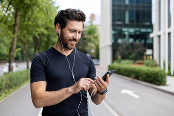 Close-up photo of a young happy male athlete standing on a city street wearing headphones and using a mobile phone