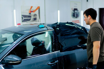 a car mechanic carefully sticks a protective tinted film on a car glass at a detailing station