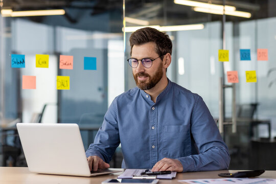 Successful and self-confident young businessman working concentratedly in the office behind a laptop. Sitting at the desk smiling, checking data and accounts, talking on a video call - Powered by Adobe