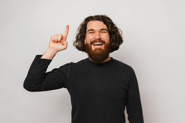 Ecstatic handsome bearded man with long hair points up with his index finger. Studio portrait over grey background.