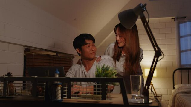 Young asian man overworking at home using laptop computer at night in bedroom. His Asian girlfriend in pajamas stands and gives him a massage, encouraging him to finish his hastily completed work.