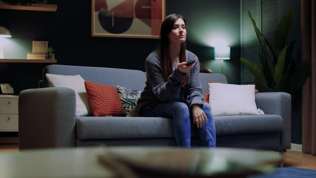 Young woman enjoys relaxing on comfortable sofa at home, watching television sitcom. Smiling lady listening to romantic soap opera or reality program on TV while sitting on couch in living room.